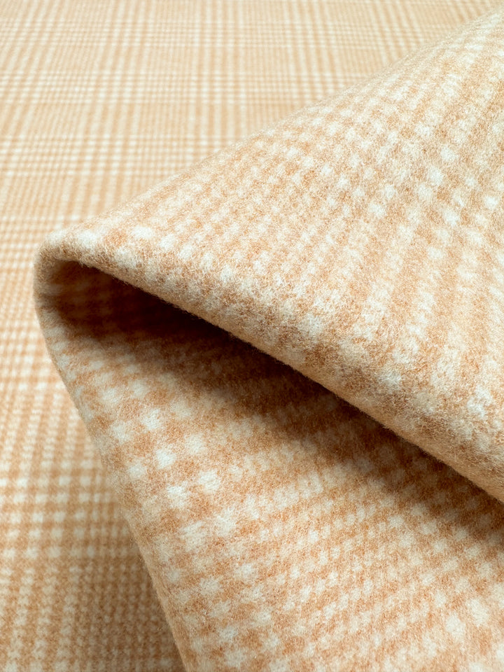 A close-up of folded camel-colored Wool Flannel - Mandarin - 148cm from Super Cheap Fabrics with a white plaid pattern. The texture appears soft and cozy, suggesting it might be used for garments like jackets, scarves, or blankets. The heavy weight fabric is neatly folded, showcasing the detailed checkered design perfect for winter wear.