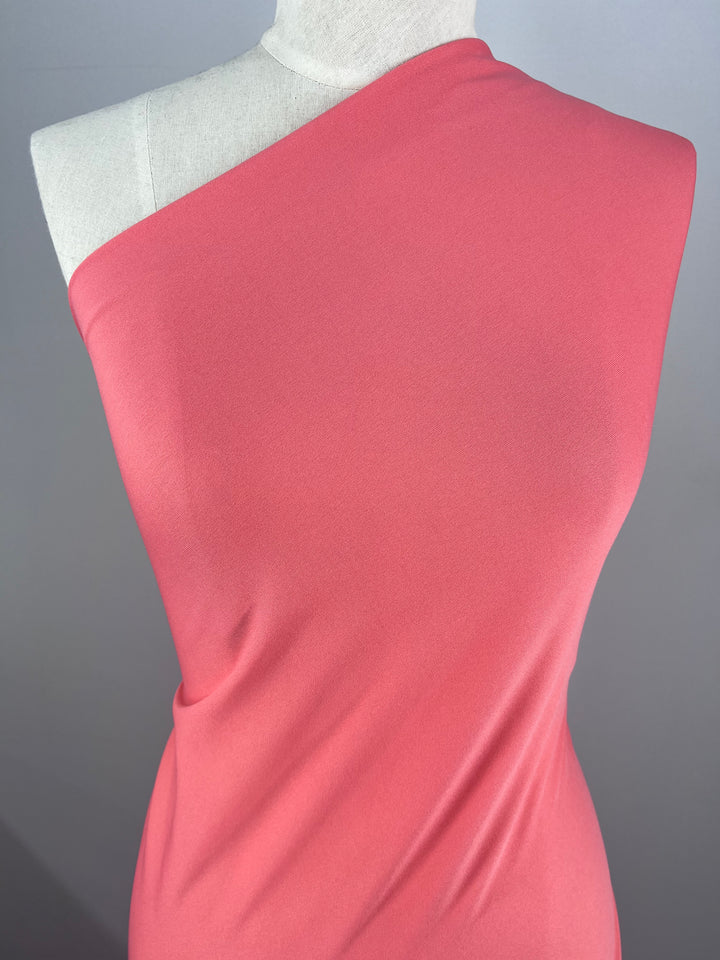 A mannequin dons the Super Cheap Fabrics Interlock in Peach Blossom, a one-shoulder coral pink polyester fabric, resting against a neutral gray backdrop. Its lightweight, smooth texture suggests a form-fitting elegance like spring's delicate blooms.