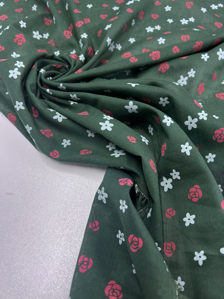 The Linen Voile - Roses by Super Cheap Fabrics, featuring small pink roses and white flowers on green linen, is neatly gathered in the center to form folds. The lightweight fabric rests on a light-colored surface.