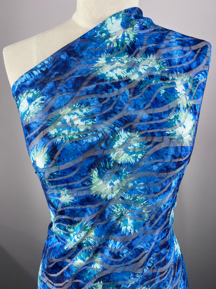 The Designer Textured Mesh - Ursula by Super Cheap Fabrics is draped over a mannequin, featuring a vibrant blue and green floral pattern with swirling lines. Its one-shoulder polyester design and lightweight texture give it an airy, flowing appearance.