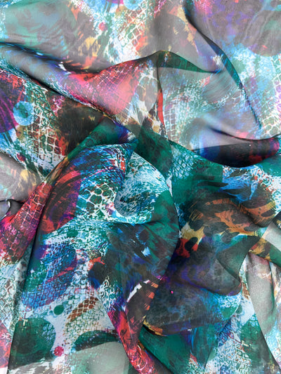 A close-up of Super Cheap Fabrics' Printed Chiffon - Petunia reveals vibrant, multi-patterned polyester fabric with swirling abstract designs in shades of green, blue, red, and white. Lightweight chiffon forms textured folds and layers ideal for creative sewing projects. Width: 150cm.