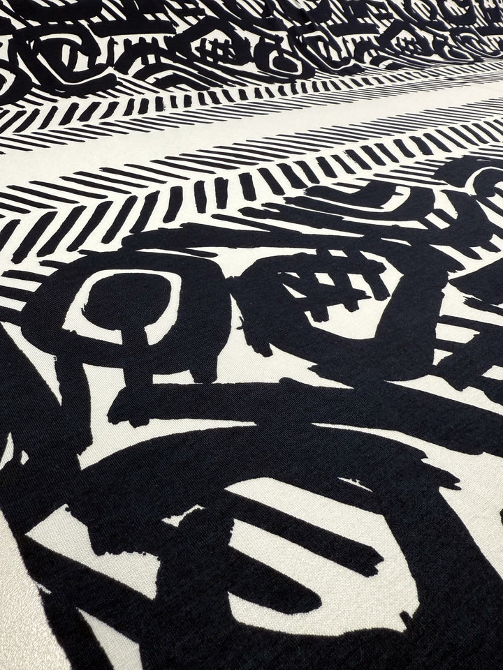 The Viscose Knit - Tribal Brushstroke by Super Cheap Fabrics features bold black abstract shapes and brushstroke patterns on cream, blending geometric lines with organic forms in a dynamic, breathable 142cm-wide summer fabric.