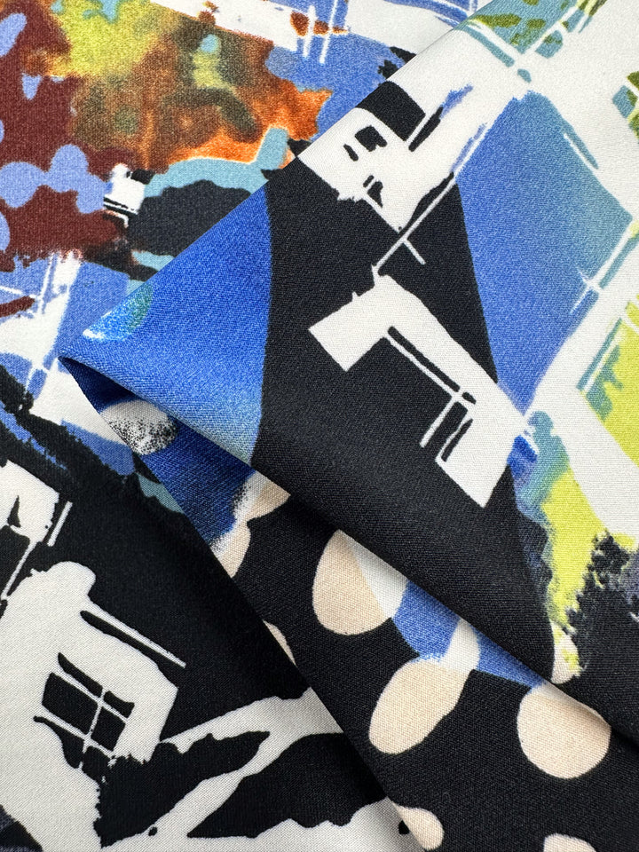 A close-up image of folded, medium-weight fabric featuring an abstract design with various colors including black, blue, white, and bits of green and brown. The Super Cheap Fabrics Printed Lycra - Tonal - 150cm pattern includes shapes resembling buildings and large white dots on a black background.