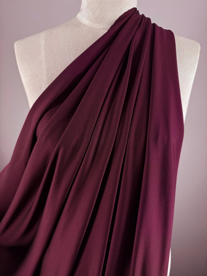 A mannequin displays Super Cheap Fabrics' Nylon Lycra - Tawny Port (154cm) arranged diagonally across the torso, set against a soft mauve background. The fabric shows gentle folds and a smooth, stretchy texture.