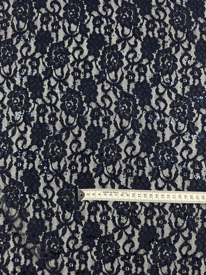 A close-up image of the lightweight Evening Lace - Night Sky from Super Cheap Fabrics, showcases an intricate floral pattern over a beige background. The design features small, shiny sequins scattered throughout. A white measuring tape is placed across the sheer black 100% polyester fabric, measuring up to 30 centimeters to show the scale.