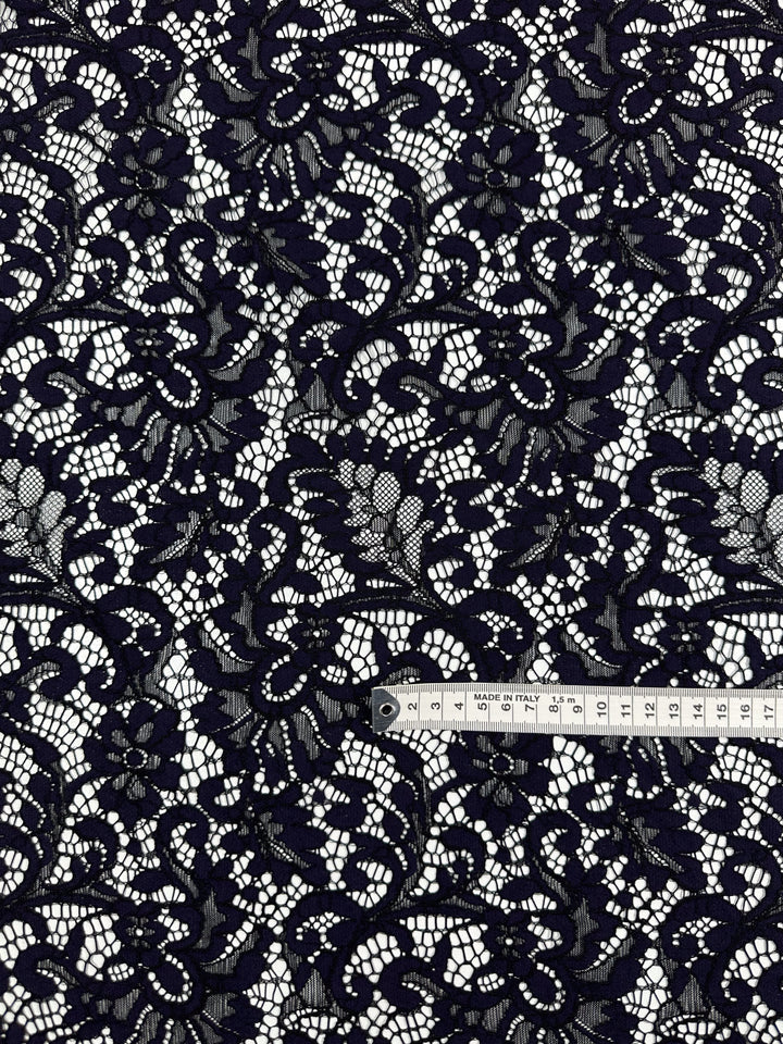 A close-up of Super Cheap Fabrics' Evening Lace - Letita - 148cm, a dark navy lace fabric featuring an intricate floral pattern. A white measuring tape with black markings lies on the fabric, showing both inches and centimeters for scale. The lightweight fabric's sheer quality and the white background beautifully highlight the lace details.