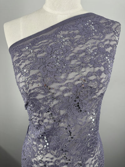 The dress form showcases a single piece of Super Cheap Fabrics' "Evening Lace - Queen - 140cm" in sheer purple. This lightweight fabric boasts an intricate floral pattern, adorned with small, shiny sequins scattered throughout. The plain gray background accentuates the texture and details of the lace beautifully.