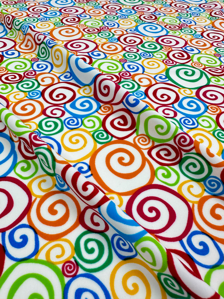 Close-up of Printed Nylon Lycra - Swirly - 155cm by Super Cheap Fabrics. The fabric showcases a vibrant, psychedelic pattern with swirling spirals in blue, red, orange, green, and yellow against a white background. This high-quality print accentuates every vivid detail on the soft material, making it suitable for various uses and ensuring a lively and dynamic visual effect.