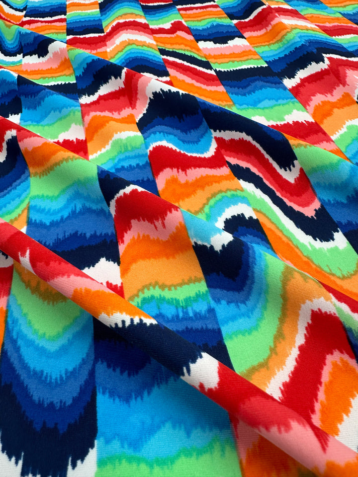 The Printed Nylon Lycra - Fuzz & Waves by Super Cheap Fabrics features a vibrant abstract pattern with wavy stripes in vivid red, blue, green, orange, and white. Its high-quality print enhances the slightly folded texture on this versatile 152cm fabric.
