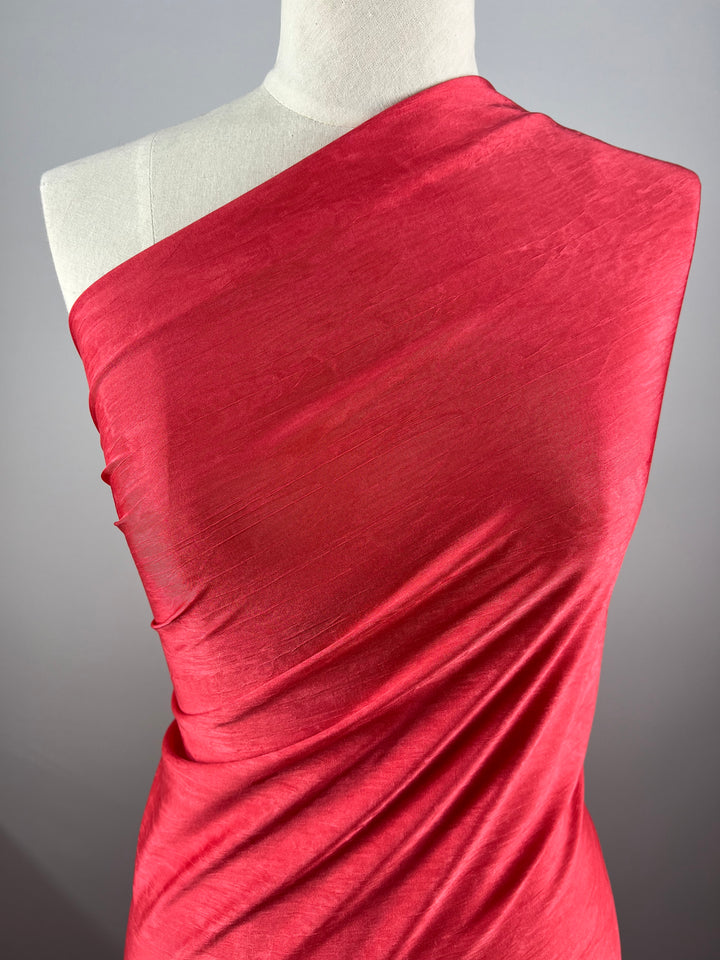 A mannequin displays Super Cheap Fabrics' Shimmer Knit - Hibiscus, a vibrant red, off-the-shoulder polyester fabric showcasing its smooth texture and elegant folds against a plain gray background.