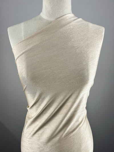 A close-up image of a mannequin draped in a light beige, one-shoulder, textured fabric made of Polyester Spandex. The Shimmer Knit - Nude - 150cm by Super Cheap Fabrics's subtle sheen enhances its elegant look. The lightweight material flows gracefully against the plain gray background, highlighting the nude colour and draped design.