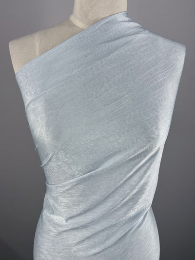 A sky-colored, shimmery Polyester fabric draped over a mannequin. The lightweight fabric is arranged to cover the mannequin from one shoulder down to the waist, showcasing its smooth texture and subtle sheen. The background is plain gray, highlighting the fabric's color and drape. Product Name: Shimmer Knit - Sky - 150cm Brand Name: Super Cheap Fabrics