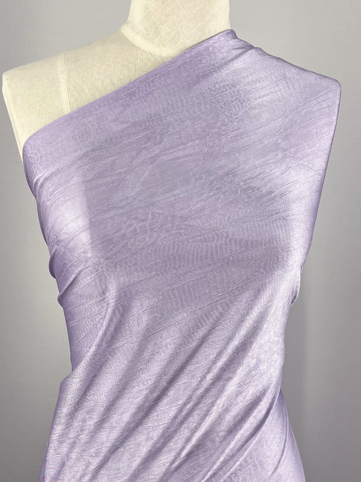 A mannequin draped with Super Cheap Fabrics' Shimmer Knit in Lilac, featuring a single-shoulder wrap made from the lightweight, subtly iridescent 150cm fabric with a faint, intricate pattern. The elegant appearance of the fabric is enhanced against the neutral background and perfectly paired with loose pants in matching lilac.