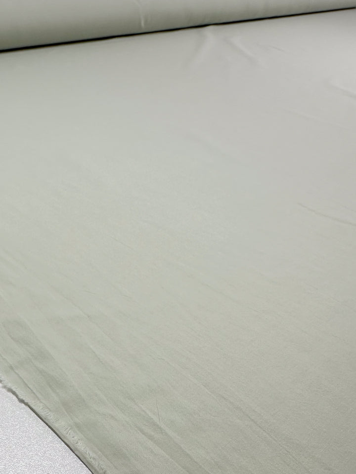 A roll of "Plain Cotton - Fairest Jade - 152cm" by Super Cheap Fabrics is partially unrolled, revealing a light grey fabric with a smooth, even texture and soft appearance.