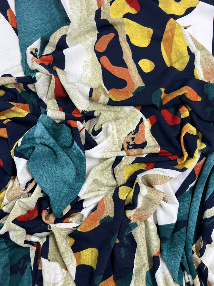 A close-up of draped Super Cheap Fabrics' Printed Lycra - Melt - 150cm fabric featuring an abstract pattern with swirls of navy blue, mustard yellow, red, and forest green. The medium weight fabric appears soft and gathered in folds, creating a textured and dynamic visual effect against the teal backing.