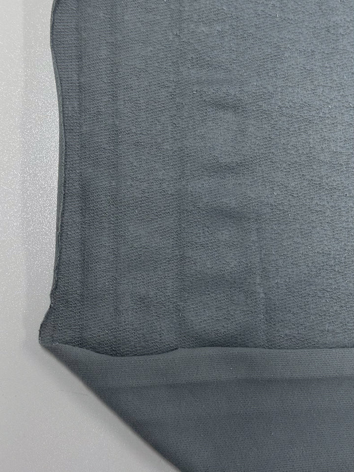 Close-up of the corner of a gray fabric, likely a textile sample. The material appears opaque with a flat texture, and its edges are slightly rough cut. Made from high-quality cotton, this "Brushed Fleece - Monument - 175cm" by Super Cheap Fabrics lies on a light-colored surface, suggesting it is intended for apparel or upholstery.