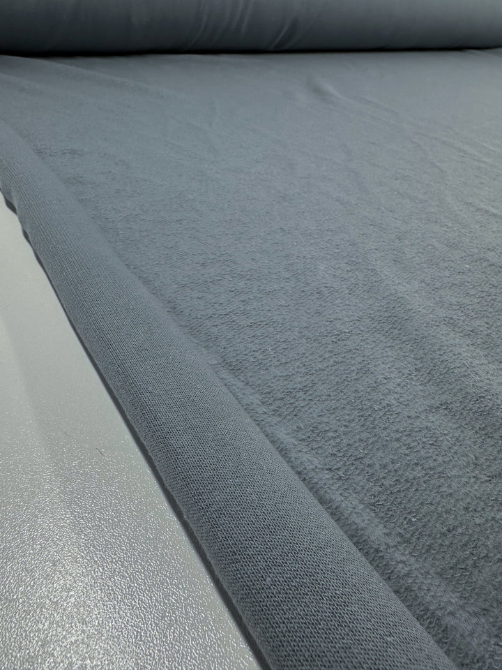 A close-up shot captures a large roll of the Brushed Fleece - Monument - 175cm from Super Cheap Fabrics, unfurled on a flat surface. The high-quality cotton fabric, which is gray in color, appears soft and subtly textured. The partially unwound roll showcases the smooth and continuous length of this luxurious material.