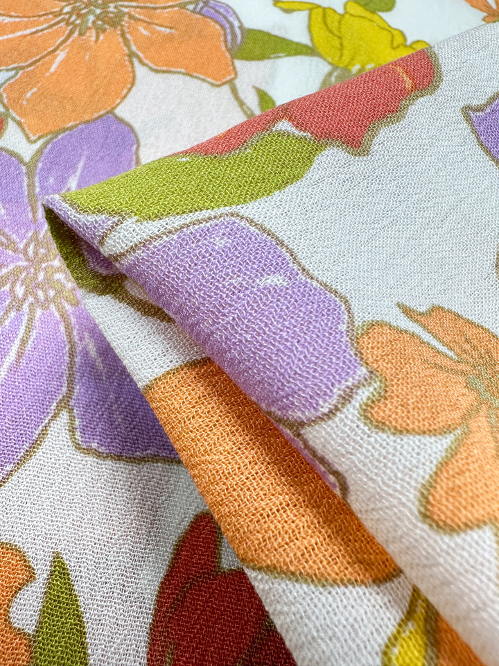 A close-up of *Soft Cotton - Illustrator - 140cm* fabric by *Super Cheap Fabrics*. This breathable summer material boasts large, vibrant flowers in shades of orange, purple, yellow, and red with green leaves set against a light background. The image captures the fabric folded to accentuate its texture and design.