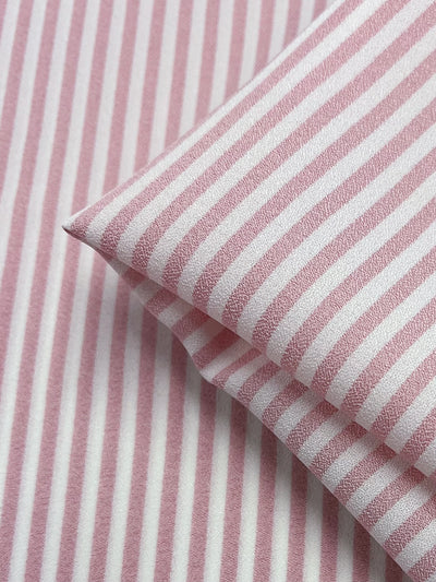 A close-up view of the Striped Crepe - Tickled Pink fabric by Super Cheap Fabrics reveals a polyester material with pink and white vertical stripes. The consistent pattern across multiple layers creates a sense of depth, enhanced by the textured appearance and neat folding. This combination exudes a tickled pink vibe that subtly conveys high fashion elegance.