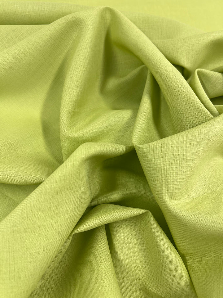 A close-up of Super Cheap Fabrics' Pure Linen in Daiquiri Green, a lightweight 100% linen fabric draped and folded to create smooth and soft contours with slight shadows. The fabric, measuring 140 cm wide, showcases a subtle woven texture visible on its surface.