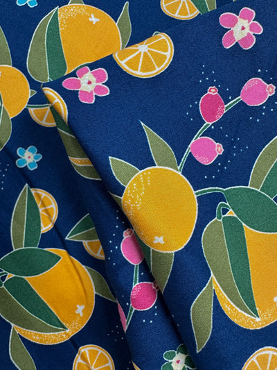 Cotton Sateen - OJ by Super Cheap Fabrics is a medium weight cotton fabric (145cm wide) with a vibrant print of yellow oranges, orange slices, pink flowers, and green leaves on a dark blue background. Pattern repeats are visible on the folded fabric.
