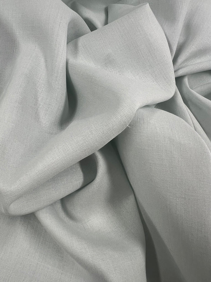 A close-up of crumpled, soft, 100% linen in a subtle Illusion Blue hue. The lightweight fabric has a smooth texture with visible weave patterns, creating gentle folds and shadows. This is the Pure Linen - Illusion Blue - 140cm by Super Cheap Fabrics.