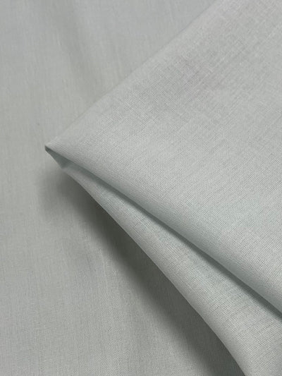 A neatly folded piece of Pure Linen - Illusion Blue - 140cm from Super Cheap Fabrics is displayed, showcasing its smooth texture and gentle folds. Made from 100% linen, the fabric has a subtle sheen, indicating a fine and soft material. The background also consists of the same Pure Linen - Illusion Blue - 140cm fabric, enhancing the uniform appearance.