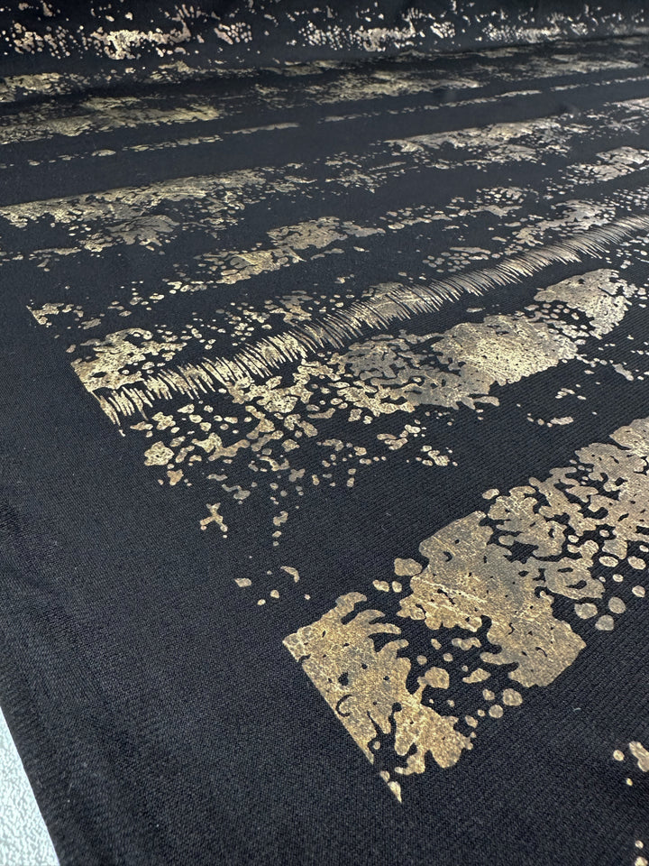 A close-up of the Evening Lycra - Rolled Gold fabric by Super Cheap Fabrics showcases its textured black Polyester/Spandex blend adorned with horizontal gold metallic patterns. This medium-weight fabric features abstract and irregular designs, offering a reflective and artistic effect across its 160cm surface.