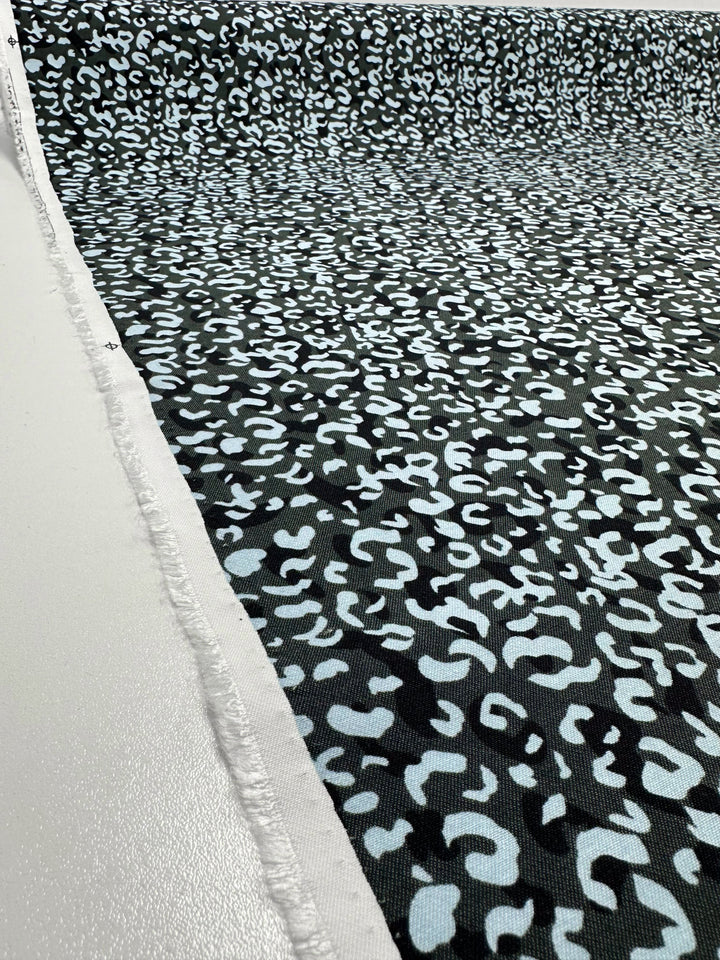 A roll of Super Cheap Fabrics' Printed Bengaline - Luana (150cm) with a black and white abstract pattern is laid out on a flat surface, showing a white selvage with frayed edges typical of woven fabric.