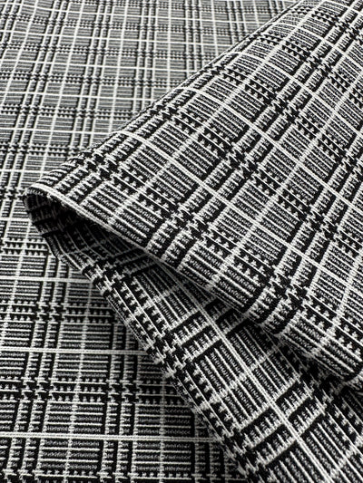 A close-up of the "Textured Bengaline - Plaidly - 150cm" from Super Cheap Fabrics reveals its black and white checkered pattern. This medium-weight fabric features intricate geometric designs that create a visually appealing and tactile surface. Some of this versatile fabric is draped over itself, showcasing the pattern from different angles.