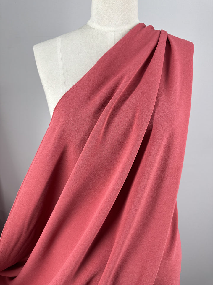 A mannequin displays "Plain Scuba Crepe - Holly Berry - 150cm" by Super Cheap Fabrics, elegantly draped over one shoulder. This medium-weight, dusty pink fabric flows gracefully with soft folds against the light gray background.