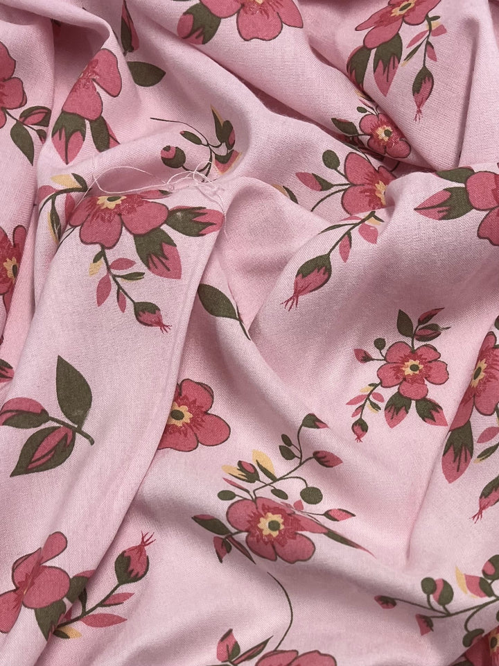 A close-up of Super Cheap Fabrics' Printed Rayon - Hippy Dais (145cm) reveals vibrant pink floral patterns with red and yellow flowers and green leaves. The versatile fabric is slightly wrinkled, forming soft folds and shadows.