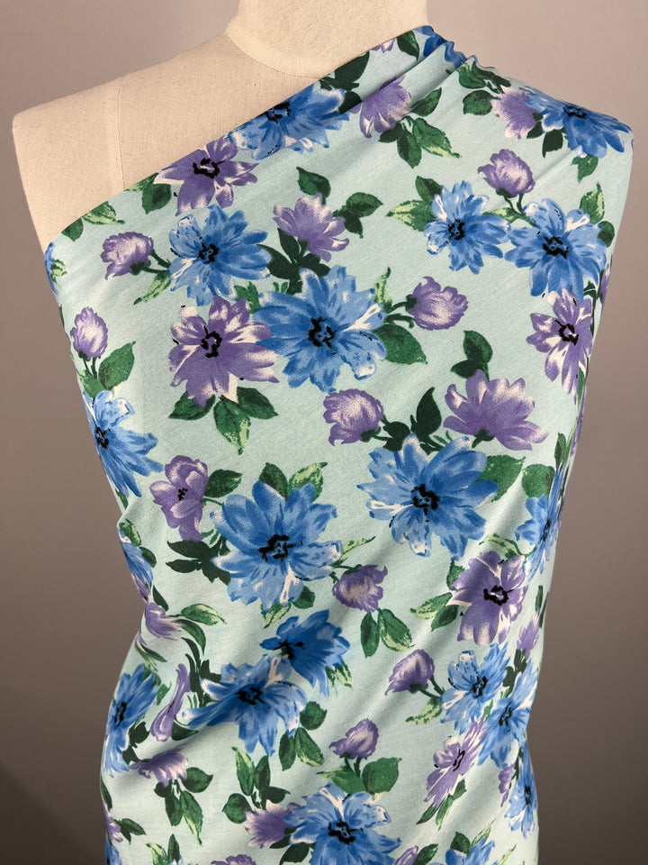 A mannequin is draped with Super Cheap Fabrics' Printed Rayon - Watercolour - 145cm, a versatile fabric featuring a vibrant floral pattern. The rayon boasts a light green background adorned with blue and purple flowers, complemented by green leaves. The display is set against a neutral backdrop.