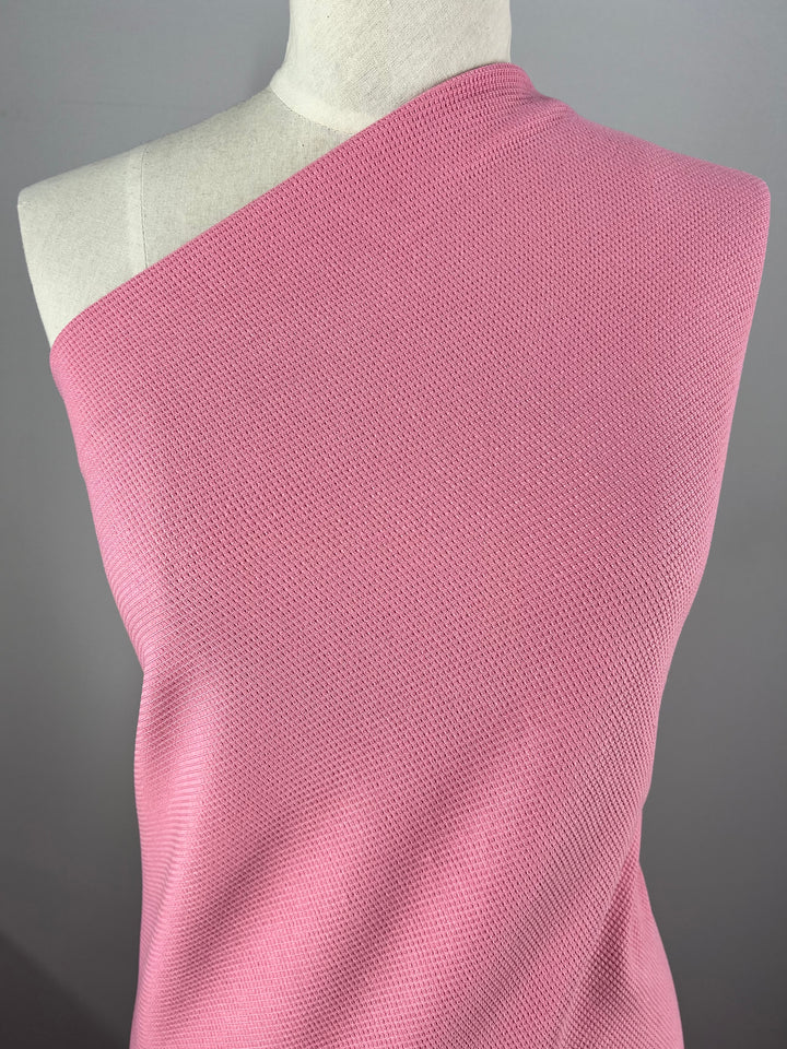 A close-up of a mannequin wearing a Super Cheap Fabrics' Waffle Knit - Peony - 170cm against a gray background. The fabric has a textured, honeycomb appearance. The left shoulder of the mannequin is bare, while the top covers the right shoulder and torso.