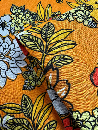 The Printed Linen Blend - Golden Garden by Super Cheap Fabrics is a bright orange medium-weight linen blend with bold yellow, white, and black floral patterns, featuring large leaves and flowers. Fabric is shown folded to highlight texture and design.