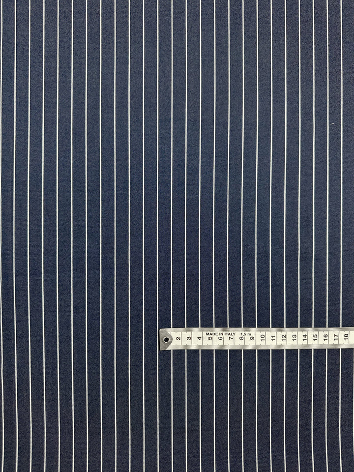 The Chambray - Sargasso Sea - 150cm by Super Cheap Fabrics features dark blue lightweight fabric with evenly spaced vertical white pinstripes. A measuring tape near the bottom shows about 18 centimeters for scale.
