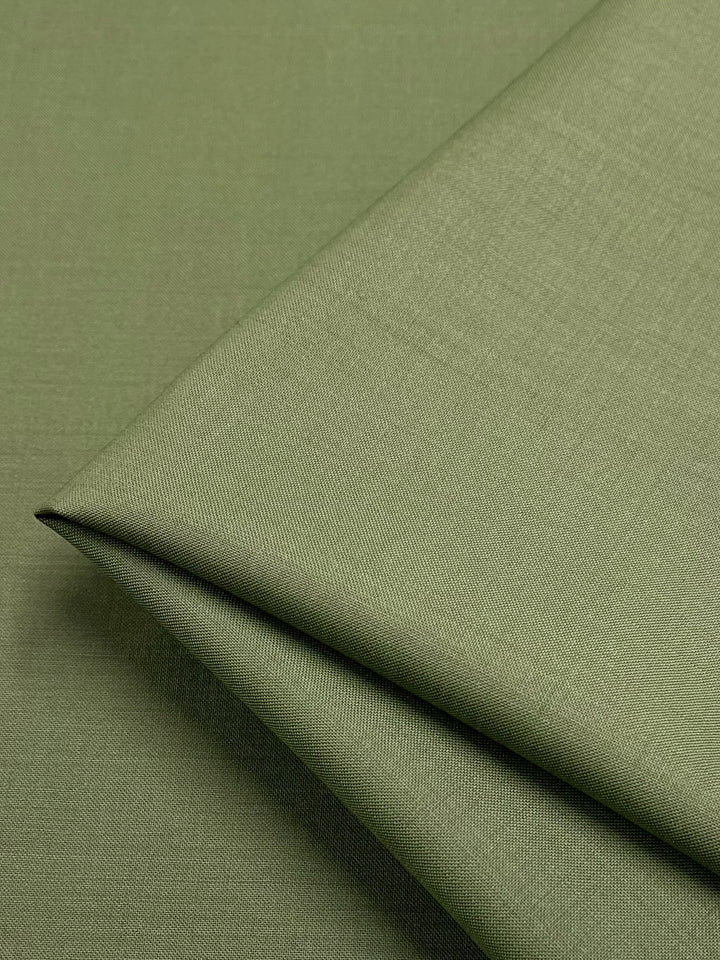 A close-up photo of a piece of Super Cheap Fabrics' Merino Wool Suiting - Hunter Green - 155cm, neatly folded. The material appears smooth and has a subtle, even texture. The fabric’s soft, muted green color is consistent throughout the image, highlighting its luxurious texture and excellent breathability.