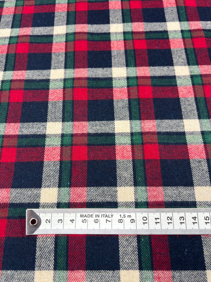 A close-up of Super Cheap Fabrics' "Suiting - Cherry Tattersal - 150cm" reveals a plaid twill fabric with red, green, black, and cream squares. A measuring tape marked in centimeters and labeled "Made in Italy" rests on this versatile material.