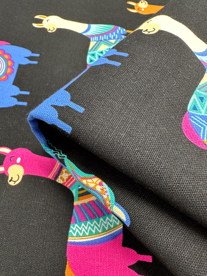 This close-up showcases the Cotton Duck - Lama Lama - 120cm fabric by Super Cheap Fabrics, featuring vibrant llama illustrations against a black background. The llamas are adorned in decorative patterns with lively pink, blue, turquoise, yellow, and orange hues, making it an ideal choice for adding a playful and artistic flair to fashion items or home décor projects.