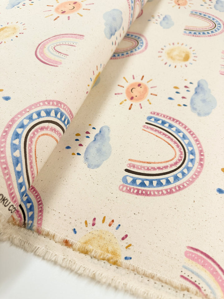 The Cotton Duck - Rainbow Natural - 112cm from Super Cheap Fabrics is a medium-weight cotton fabric that showcases a whimsical design with colorful rainbows, suns, clouds, and raindrops on a light beige background. The edges are visible, highlighting the playful pattern in soft pastel colors—ideal for home décor projects.