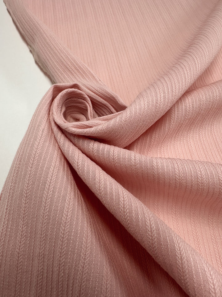 Super Cheap Fabrics' Textured Cotton - Gossamer Pink (138cm) features light pink fabric with subtle vertical stripes, loosely folded and curled at one end for a soft, elegant look on a flat surface.
