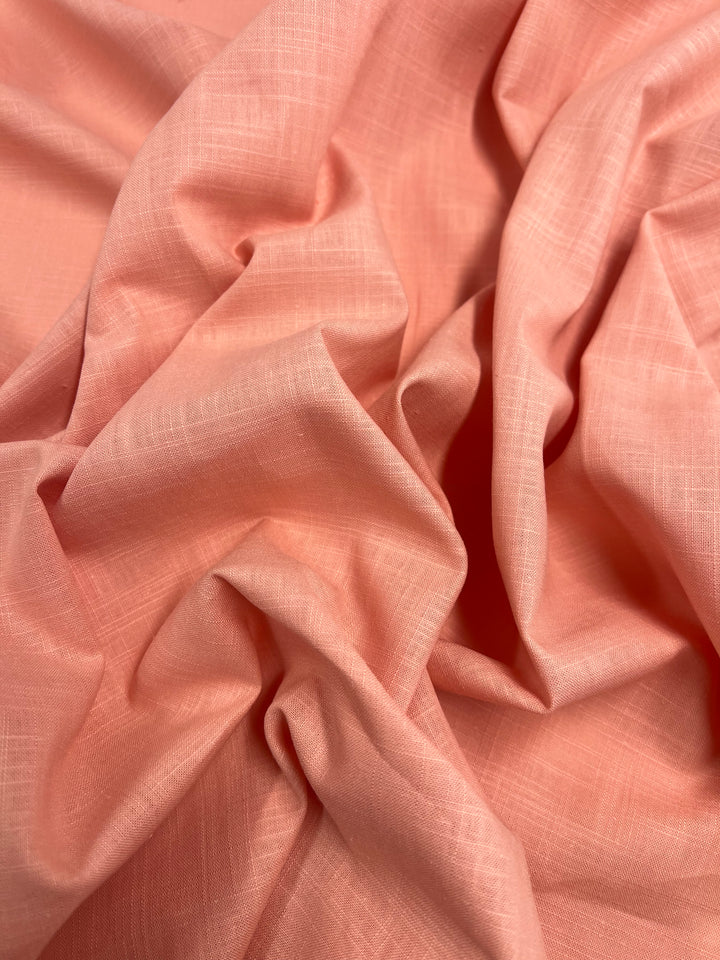 A close-up photo of soft, crumpled peach-colored fabric with visible weave texture. The Linen Blend - Coral Reef - 140cm from Super Cheap Fabrics, reminiscent of light weight linen, exhibits gentle folds and light shadows, giving it a slightly wrinkled but smooth appearance.