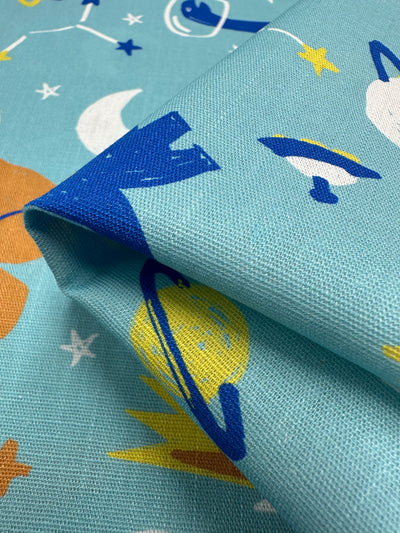 Close-up of Super Cheap Fabrics' Printed Linen - Dino Space (145cm), a blue linen with a vibrant space-themed design featuring planets, rockets, stars, and moons. Ideal for home decor or garments, the slightly folded fabric highlights texture and pattern variations.