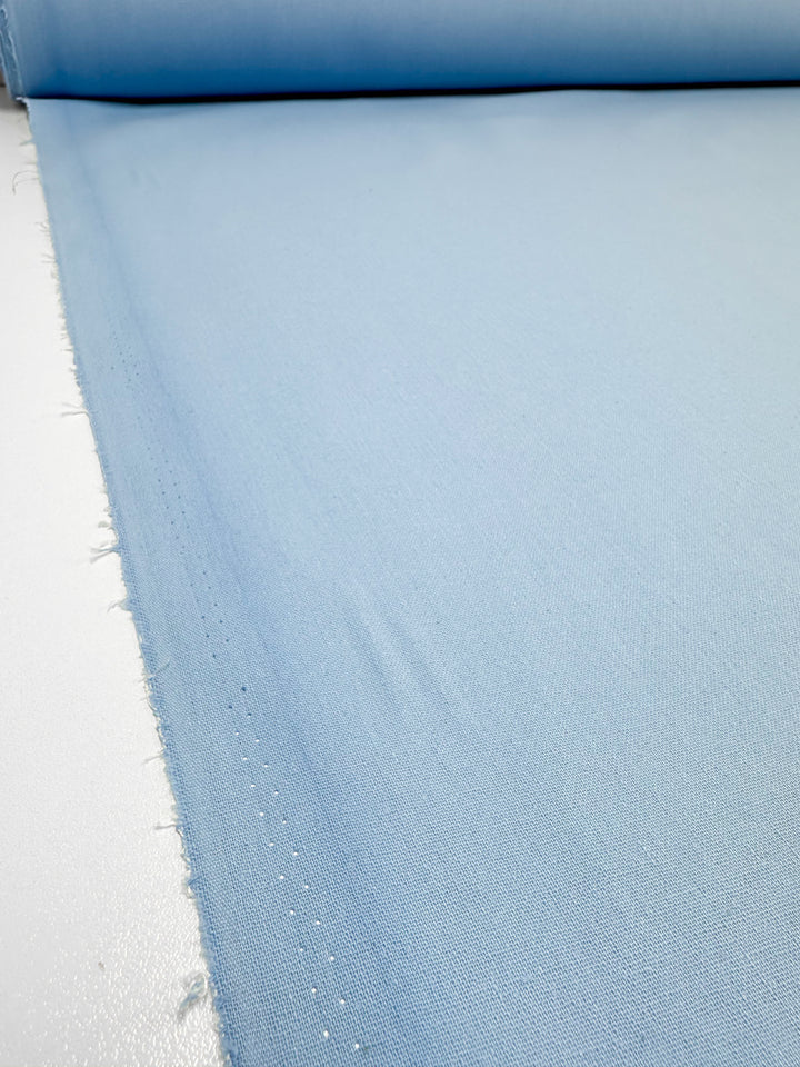 A large roll of durable Ramie Linen in "Clear Sky" light blue from Super Cheap Fabrics is spread out on a flat surface. This 150cm material has a smooth texture with slightly frayed edges, making it soft and ideal for various textile projects.