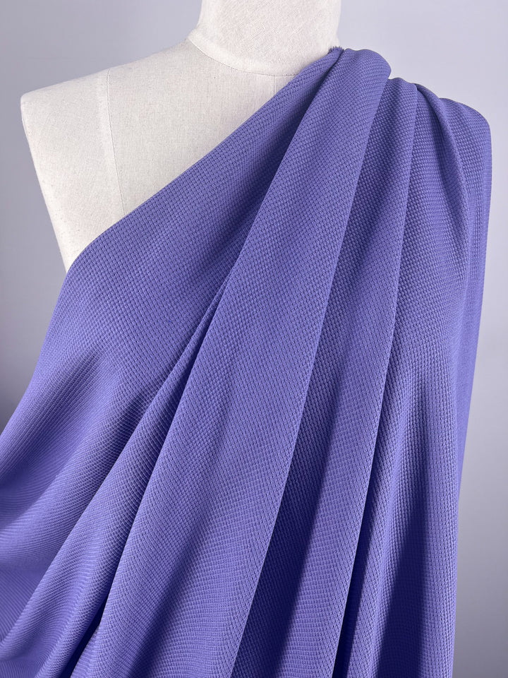 The close-up features a mannequin adorned with Super Cheap Fabrics' Waffle Knit - Sunlit Allium, a light purple fabric with a textured, three-dimensional look. Elegantly draped over the shoulder, this exquisite material covers one side of the mannequin's upper body against a plain, light backdrop.