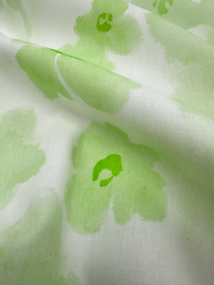 Close-up of the Super Cheap Fabrics' Printed Cotton - Lime Bloom - 150cm, featuring a white background with delicate light green floral patterns. The fabric displays a soft and slightly draped texture, accentuating the watercolor-like design of the flowers—a perfect choice for crafting clothing and household items using high-quality natural cotton fibers.