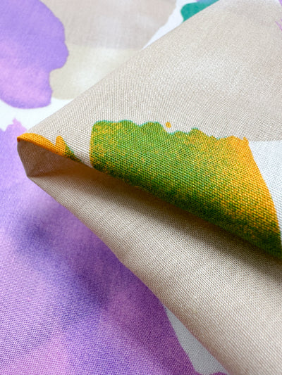 A close-up of the folded fabric "Printed Cotton - Petals - 150cm" by Super Cheap Fabrics reveals a multi-use textile. It boasts a colorful floral design with large purple flowers and green and yellow leaves on a light beige background, highlighting vibrant and delicate textures crafted from natural cotton fibers.