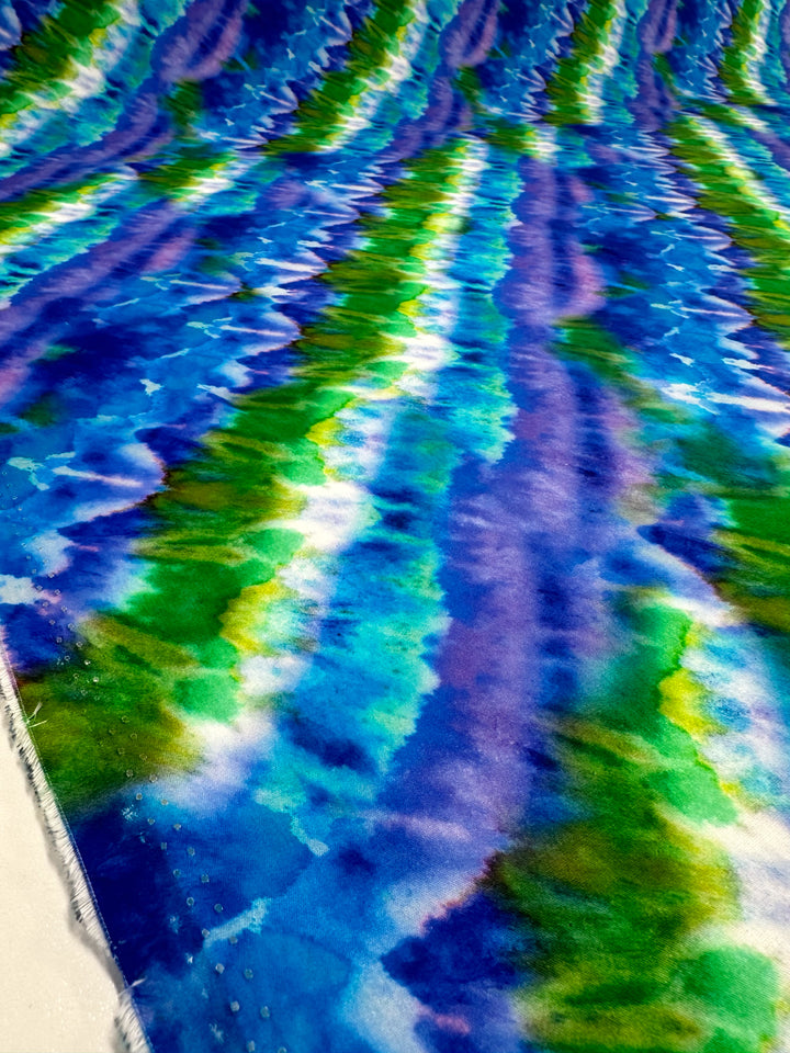 The "Printed Cotton - Lizard - 150cm" from Super Cheap Fabrics showcases a vibrant abstract pattern on lightweight fabric, featuring swirling shades of blue, green, and purple that emulate a tie-dye look with a wavy and flowing design.