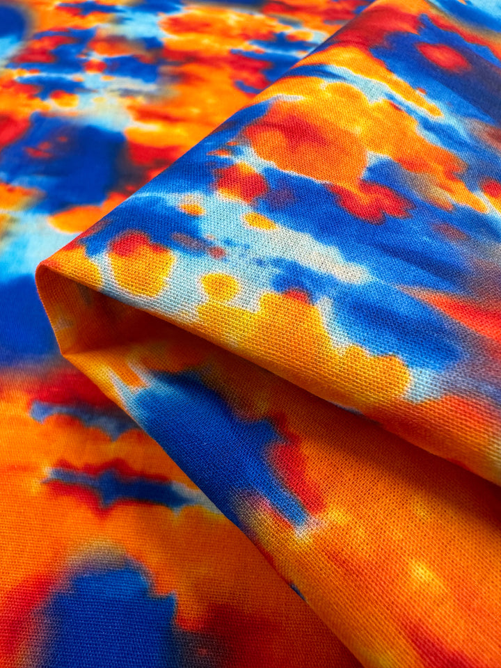 A vibrant piece of Printed Cotton - Rosch from Super Cheap Fabrics, measuring 150cm, displays a striking tie-dye pattern in shades of blue, orange, yellow, and red. The soft and natural fiber cotton is elegantly folded to highlight its bold and colorful abstract design.