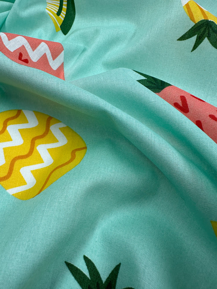 Here’s a close-up of "Printed Cotton - Pina Coalda - 150cm" by Super Cheap Fabrics, featuring a turquoise background with vibrant fruit patterns, including yellow zigzag shapes and pink triangles adorned with green leaves. Crafted from natural cotton fibers, the fabric's slightly wrinkled texture adds depth, making it ideal for clothing and household items.